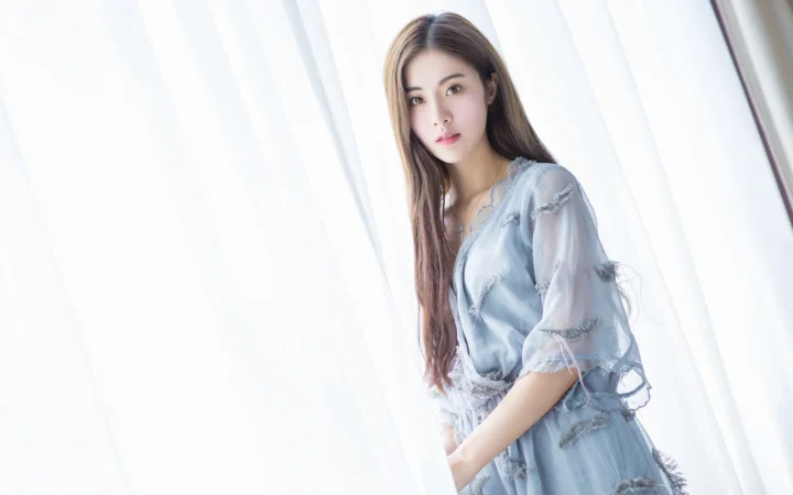 一名成年女子穿着浅蓝色服装站在白色窗帘旁。