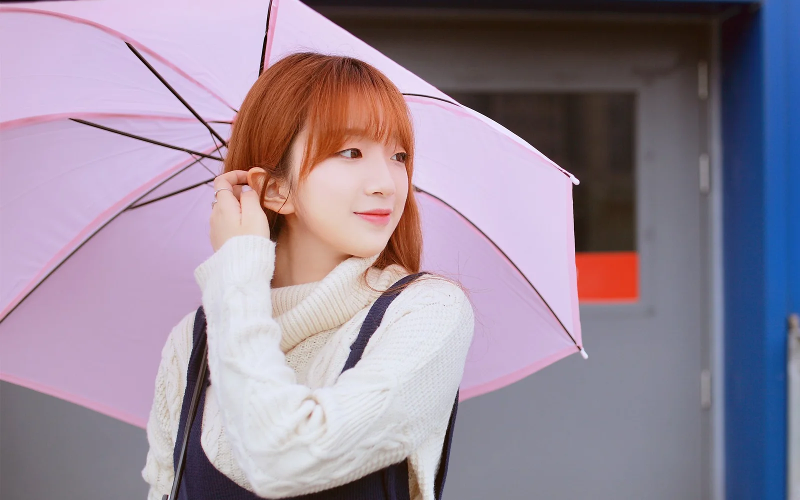 赤毛の女性がピンクの傘を持って建物のそばに立っている。
