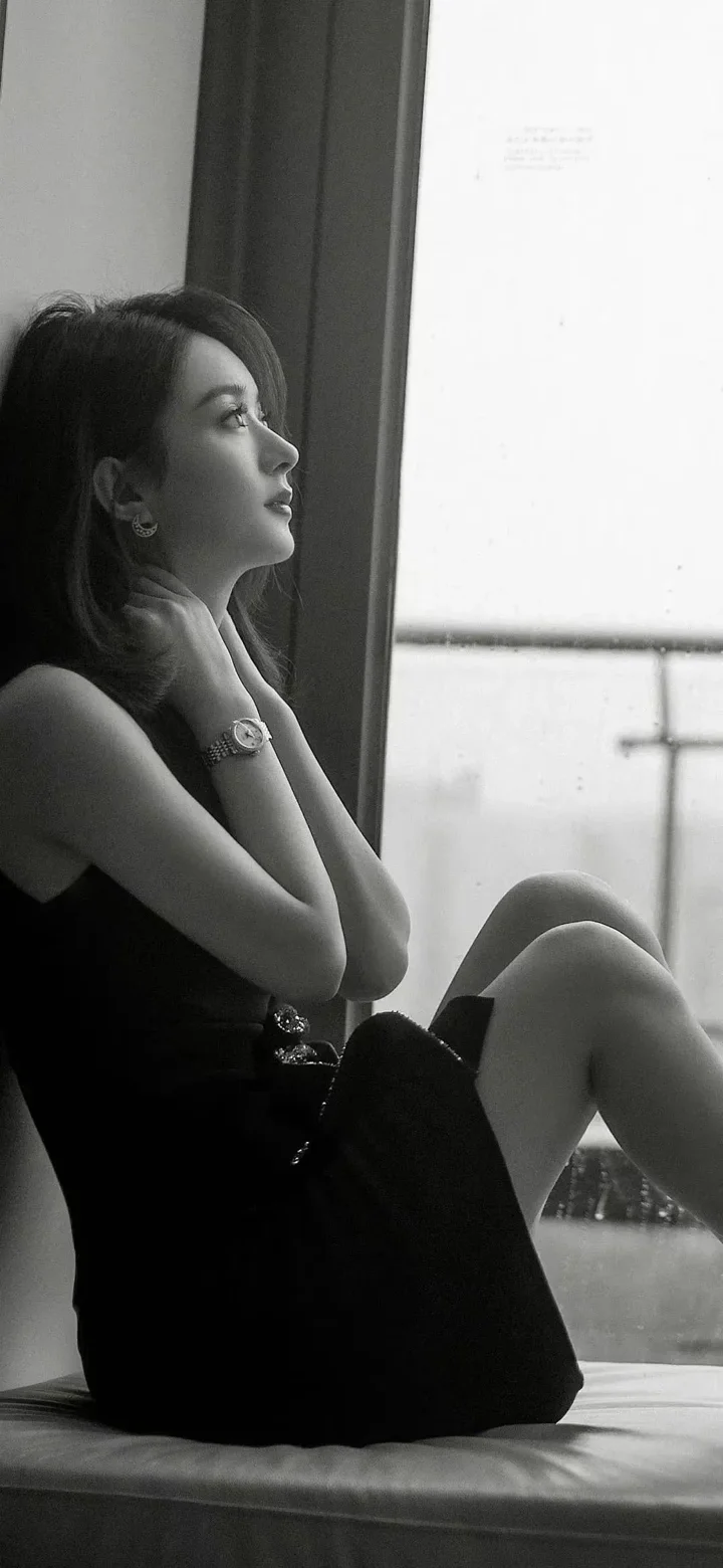 一位身穿黑色无袖连衣裙的成年女性坐在窗边，画面为黑白色调。