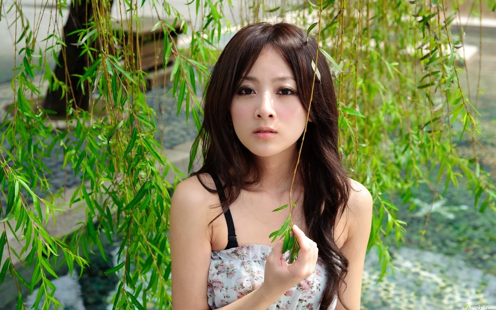 一名成年女性站在垂柳枝叶间，面向镜头微微举起手中的柳枝。