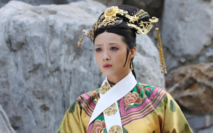 一名身穿华丽传统服饰、佩戴 ornate 头饰的成年女子站在岩石背景前。