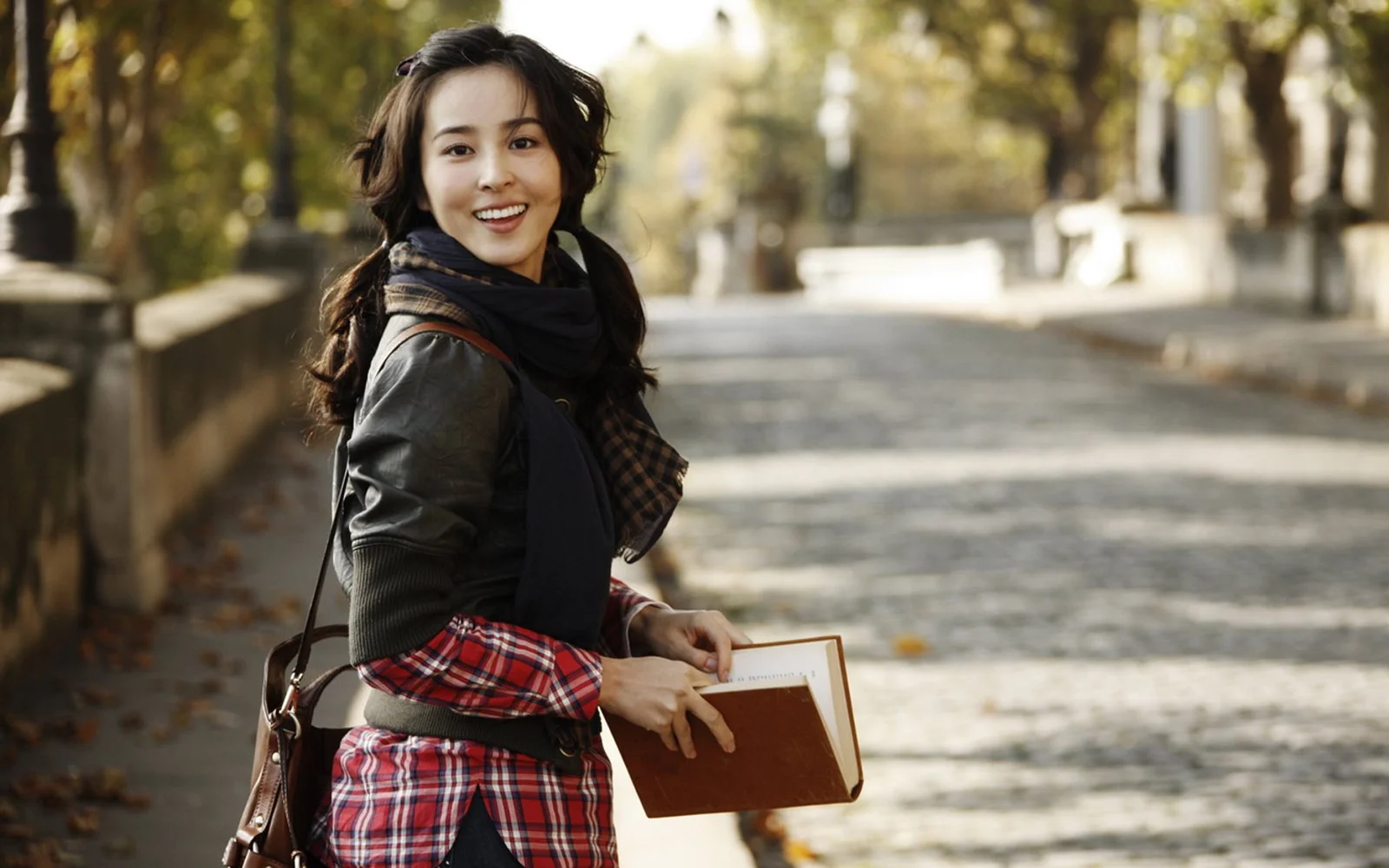 一名微笑的女子围着围巾、穿着格纹服装，手捧一本书站在林荫小径旁。