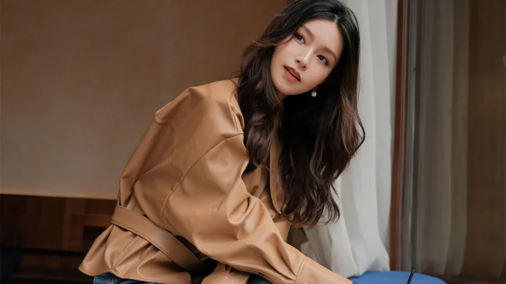 一名长发女性穿着棕色外套坐在室内窗边。