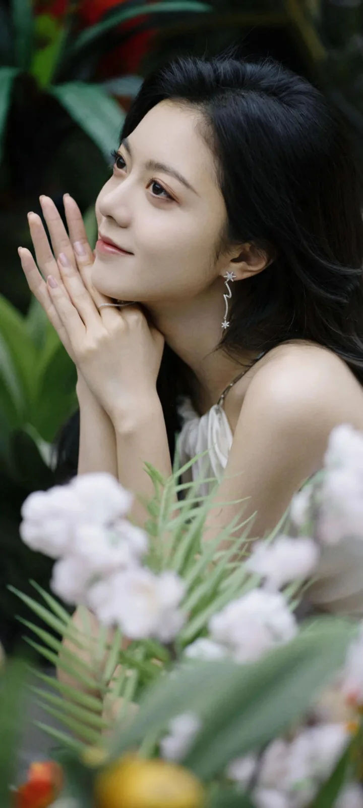 一名女性在绿叶和浅色花朵前侧脸凝望，双手轻托脸颊。