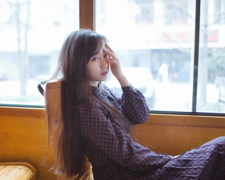 긴 머리의 여성이 차창 옆에 앉아 한 손으로 이마를 만지며 밝은 창밖을 바라보고 있다.