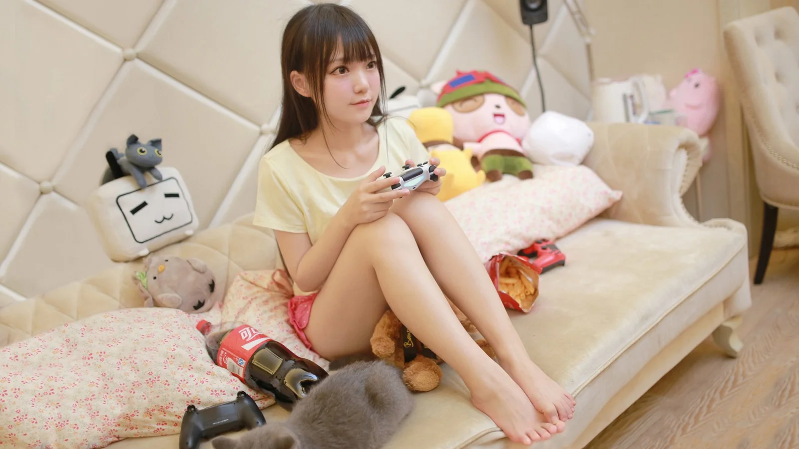 一名年輕女子坐在擺放著毛絨玩具的室內沙發上，手持遊戲手柄。