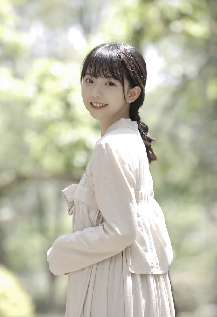 밝은 색상의 옷을 입고 포니테일을 한 여성이 초록빛 야외 배경 앞에서 미소 짓고 있다.