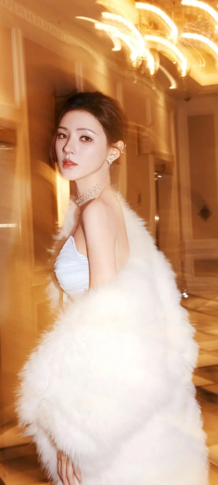 따뜻한 조명의 실내에서 흰색 퍼 숄을 두른 성인 여성이 카메라를 바라보고 있습니다.