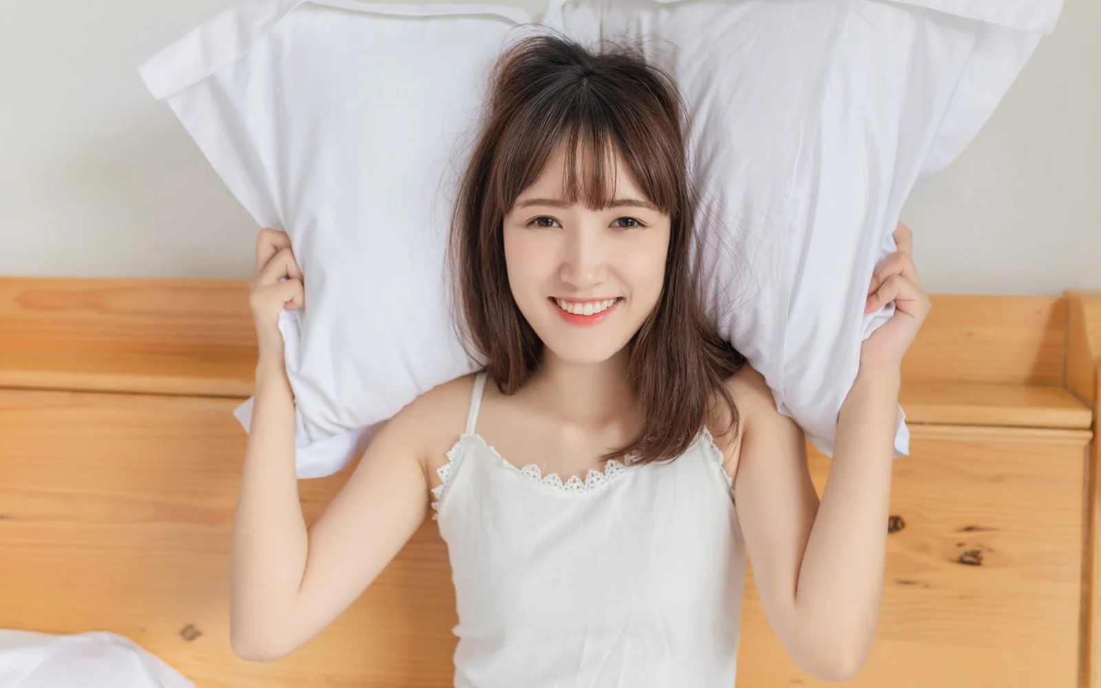 一名成年女性躺在木质床上，手持白色枕头并面带微笑。