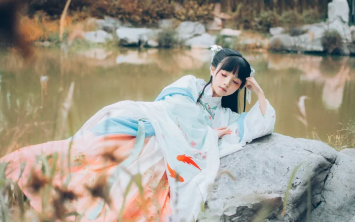 一名身穿白色传统服饰的女子倚靠在池塘边的岩石上。