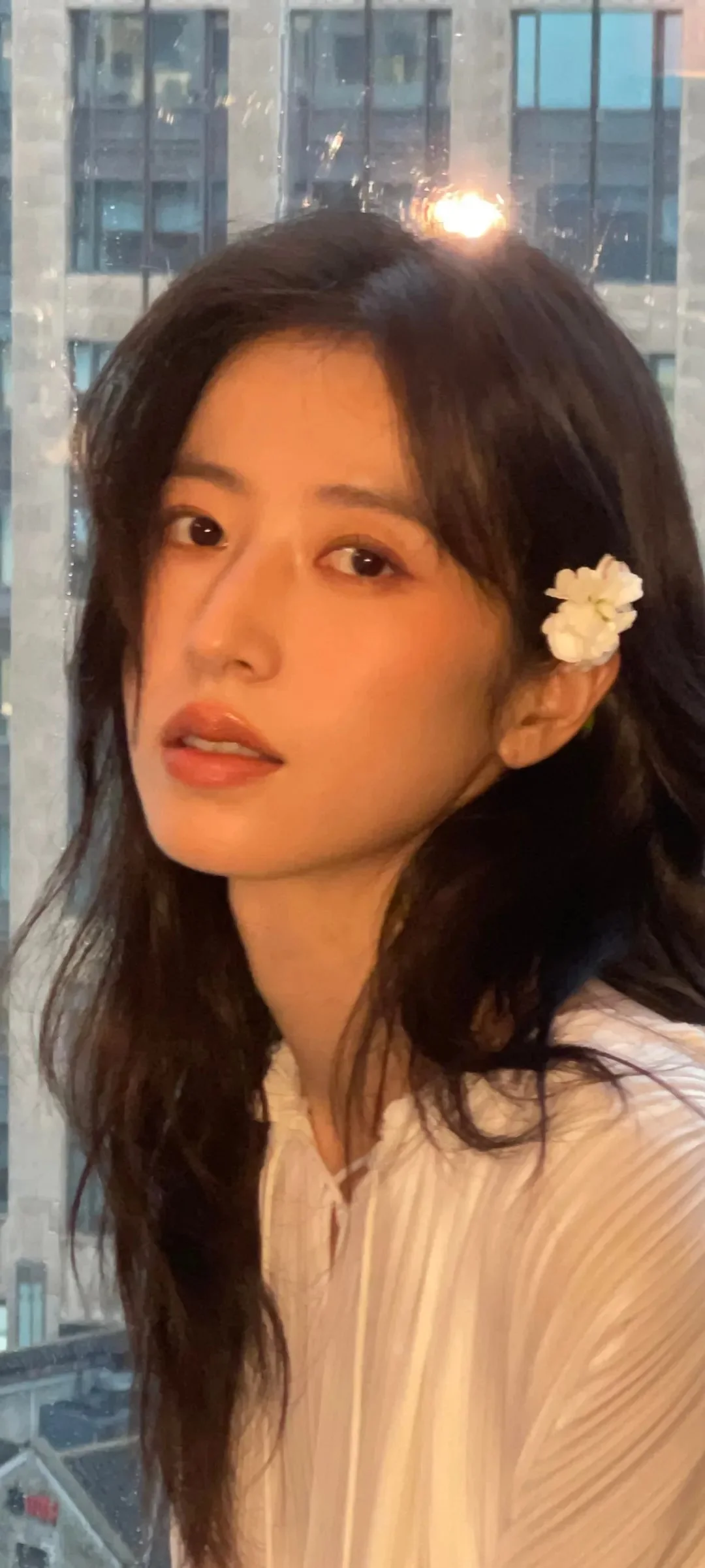 긴 검은 머리와 흰 꽃 머리 장식을 한 여성이 창가에서 카메라를 바라보고 있으며 뒤로 도시 건물이 보입니다.