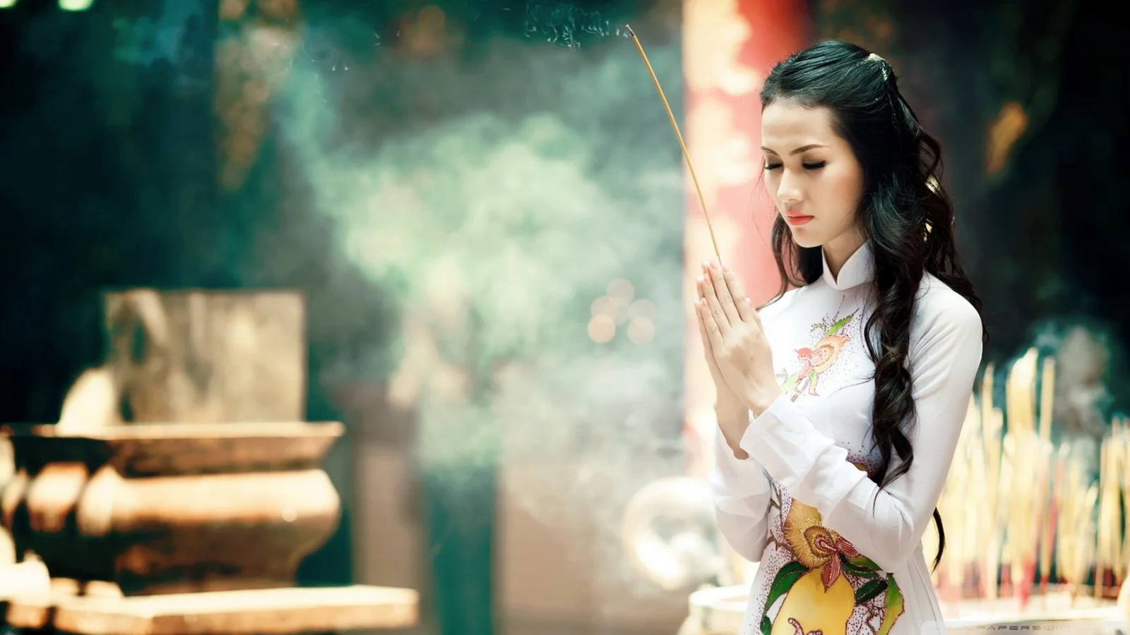 一名身穿白色服装的成年女子在香炉和烟雾前双手持香。