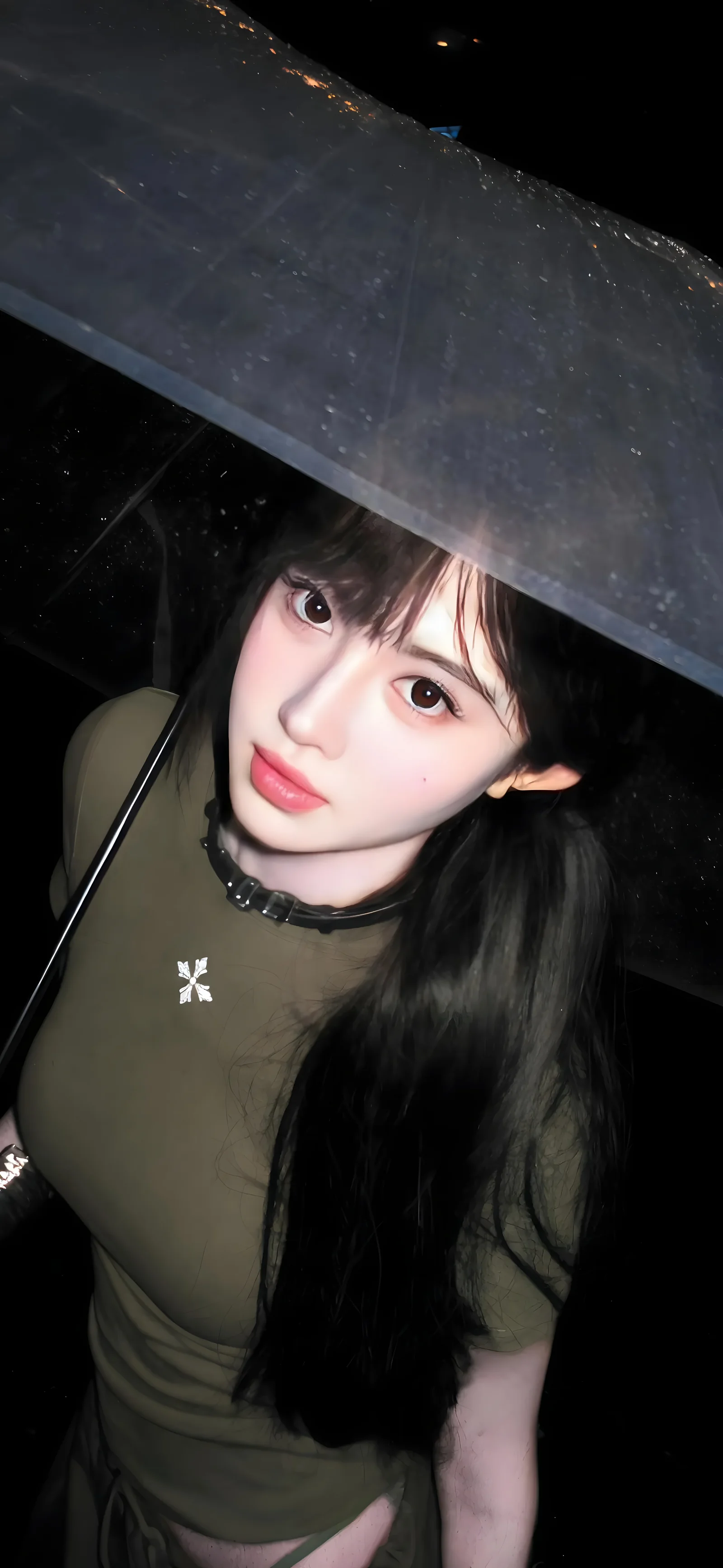 一名长发女子在黑暗背景下撑着透明雨伞，身穿橄榄绿色上衣并佩戴黑色项圈。