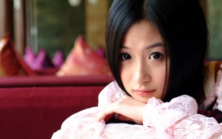 一名黑发女子身穿粉色蕾丝上衣，双臂交叠靠在沙发上面对镜头。