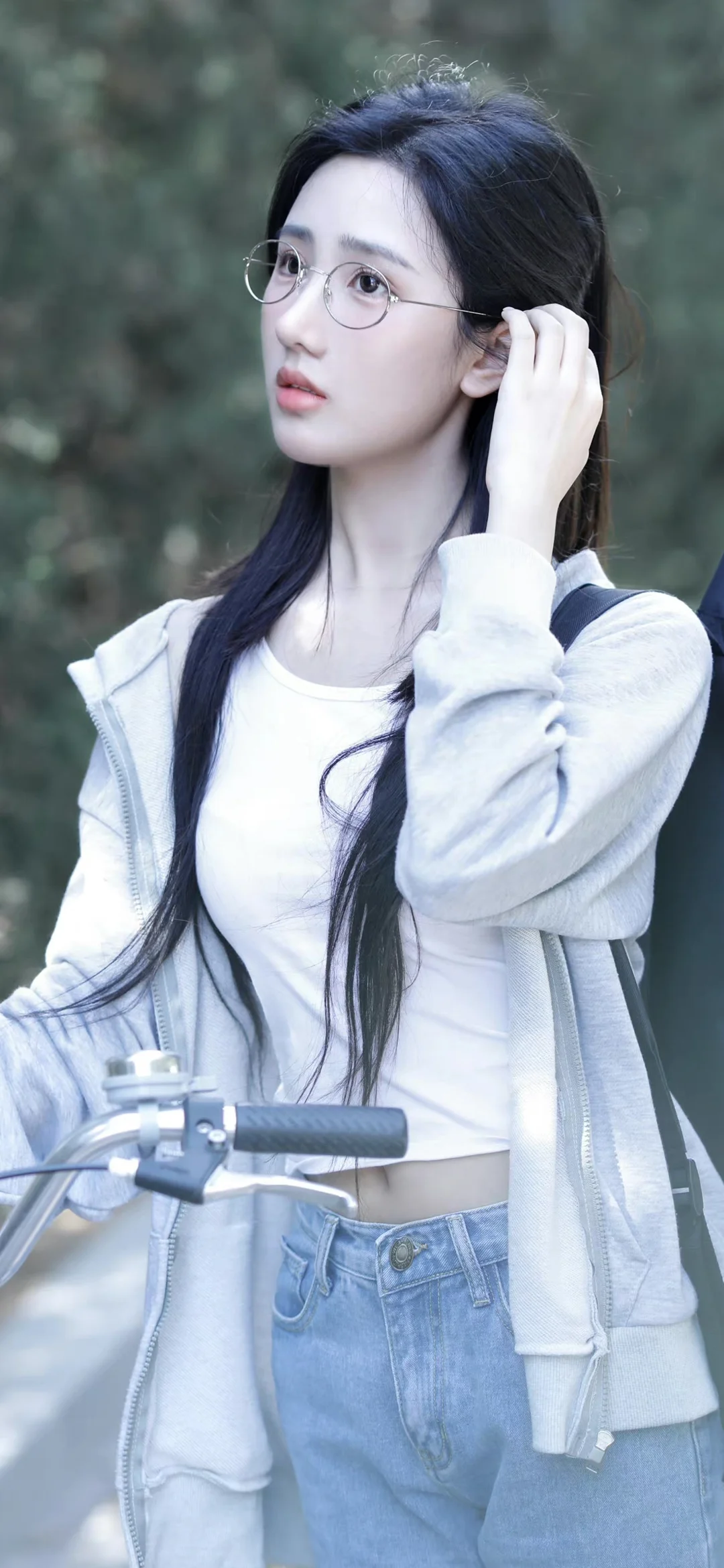 一名戴眼镜的长发女子站在自行车旁，身穿浅色连帽外套，背景为虚化的户外绿意。