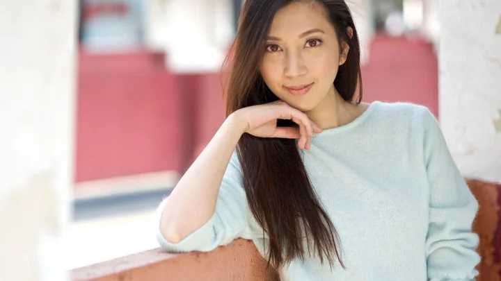 長い髪の成人女性が淡い青色のセーターを着て室内に座り、カメラを見ている。