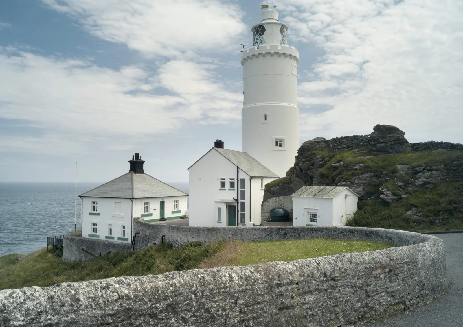 一座白色灯塔和附属建筑坐落在英国岩石海岸边，远处是海洋与多云天空。