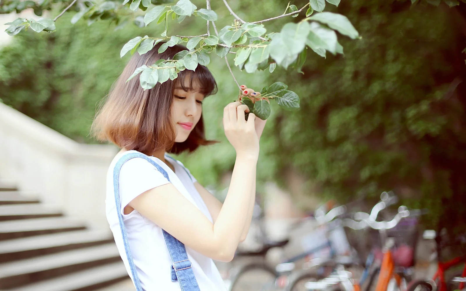 若い女性が屋外の木の枝の下に立ち、葉を手にしており、背景には自転車が見える。