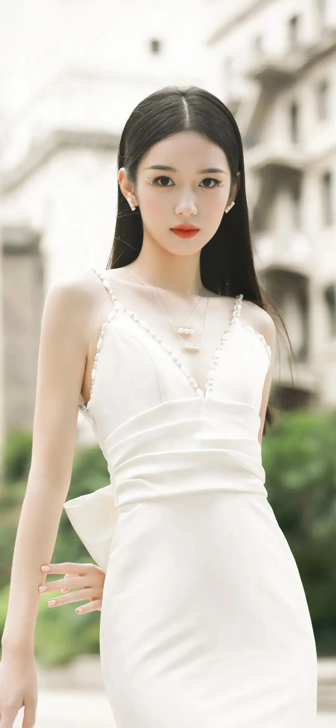 一位长发女性身穿白色连衣裙站在户外建筑背景前。