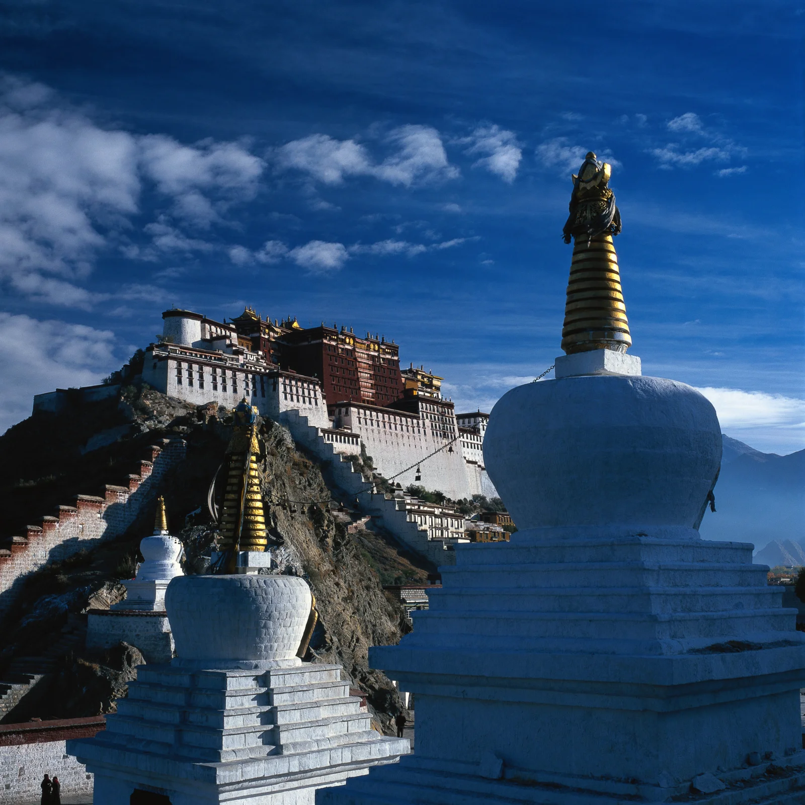 蓝天下，山坡上的宫殿建筑与前景白色佛塔映入眼帘，远处可见群山。