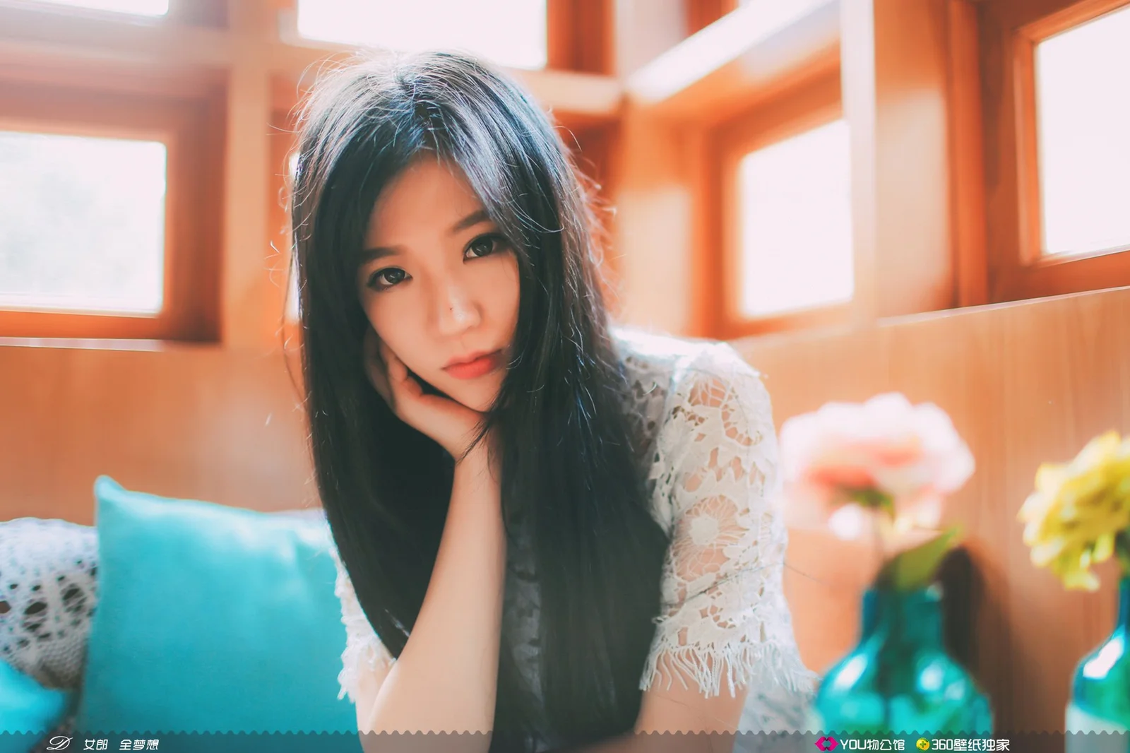 一名长发女子坐在室内窗边，以手托腮看向镜头。