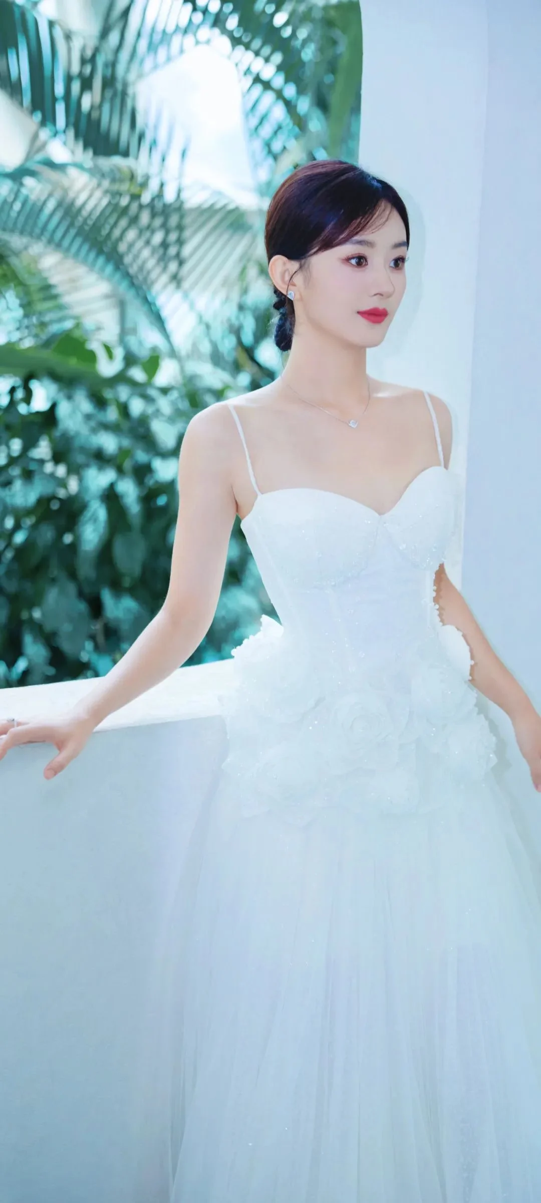 白いドレスを着た女性が、緑の植物の見える明るい室内の窓辺に立っています。