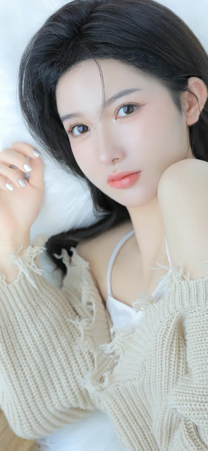 一名黑发女子穿着浅色针织衫，靠在柔软的浅色背景前看向镜头。