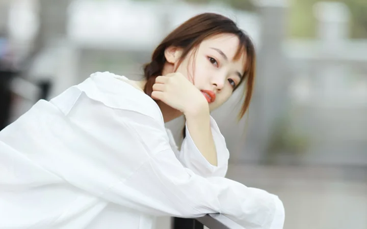 一名身穿白衬衫的女子侧身靠坐在室内，手托下巴望向镜头。