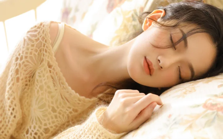 成人女性が目を閉じて枕元に横たわり、ベージュのニットを着て柔らかな暖色の光に包まれている。