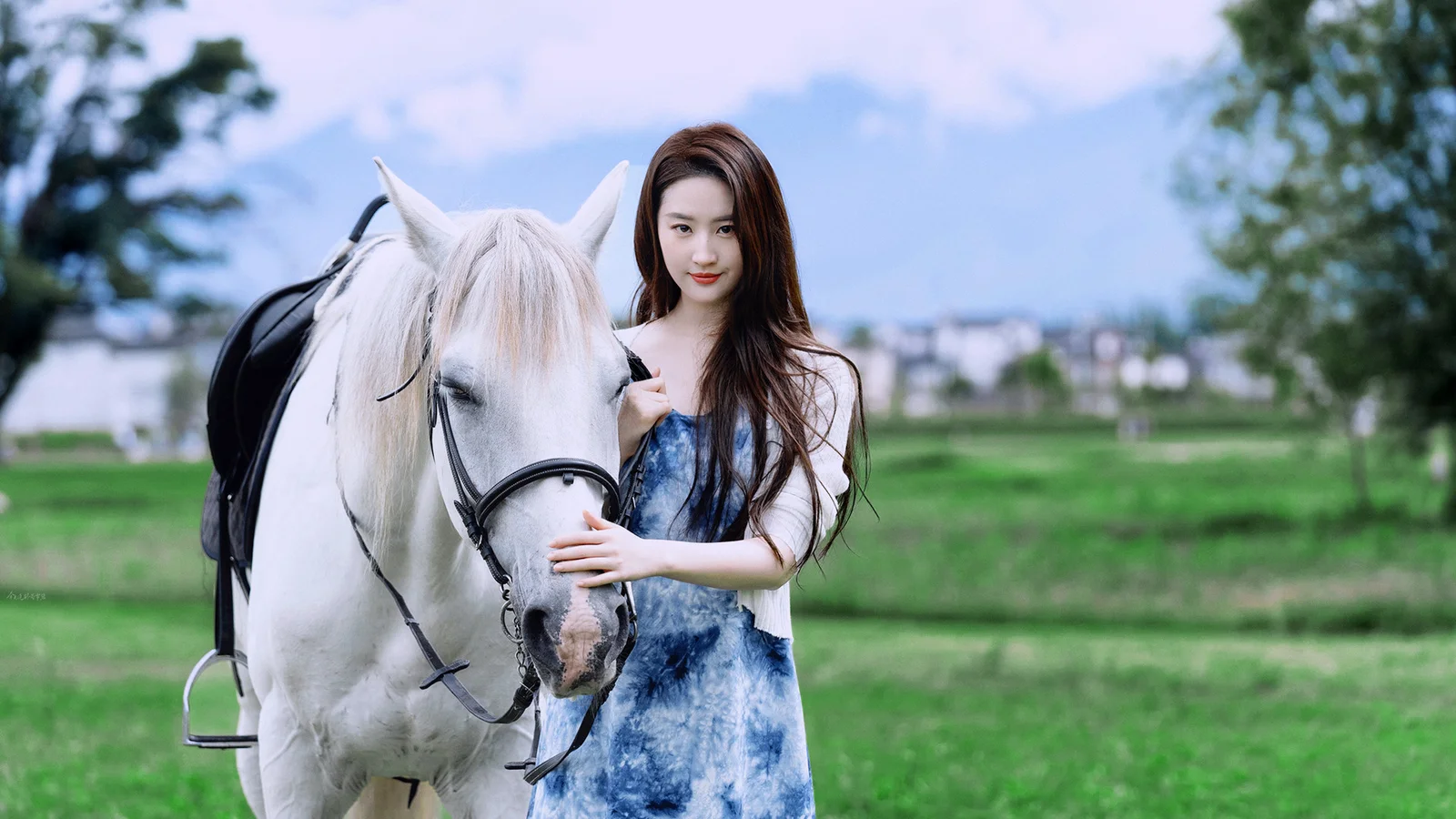 一名女子站在绿地上，身旁是一匹白马，远处可见树木和低矮建筑。