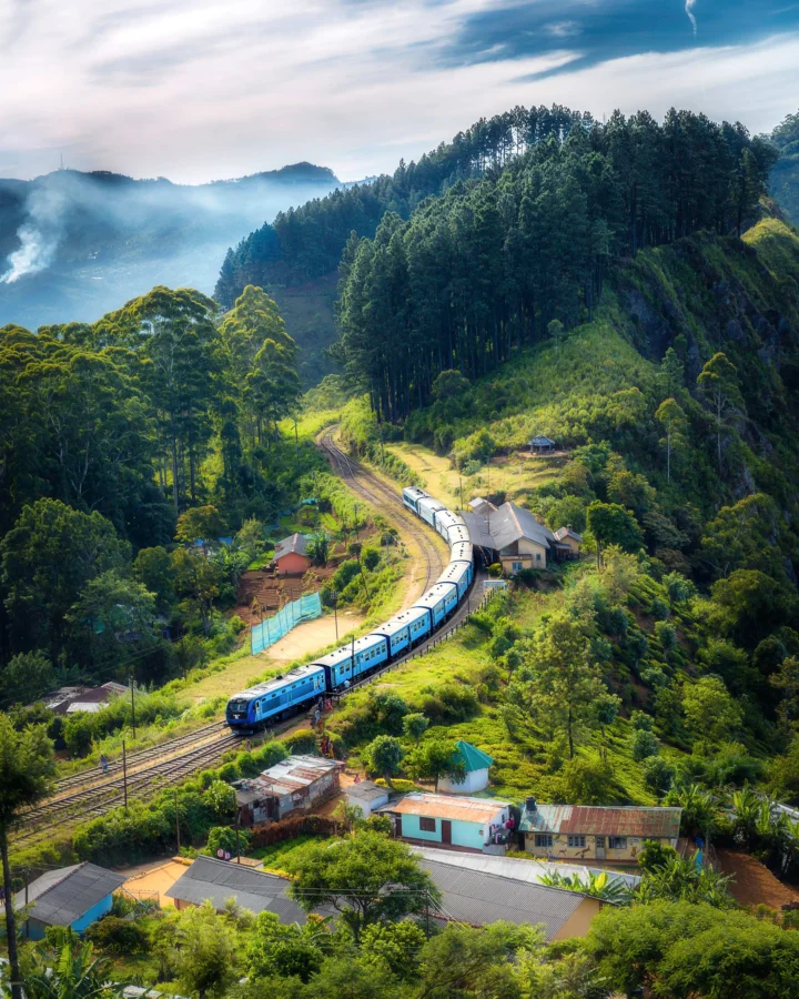 蓝色列车沿着山林间的铁路穿行，周围环绕着层叠山峦、浓密森林和山间房屋。
