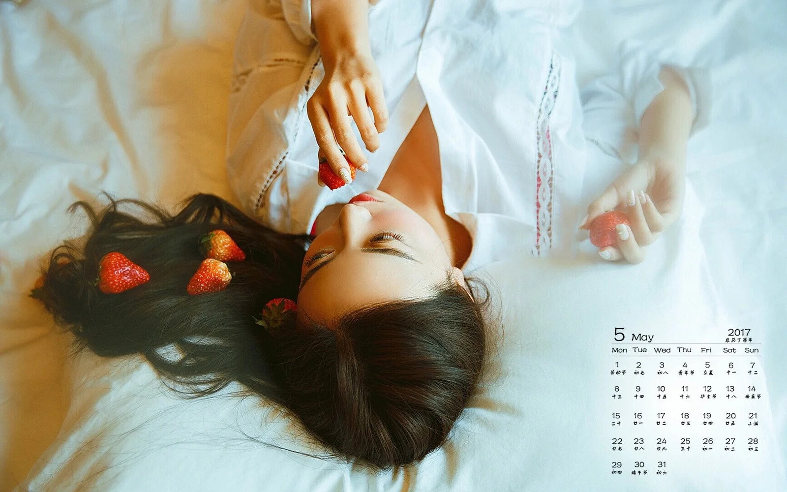 一名女子躺在白色床单上，头发旁放着几颗草莓，手中也拿着草莓。