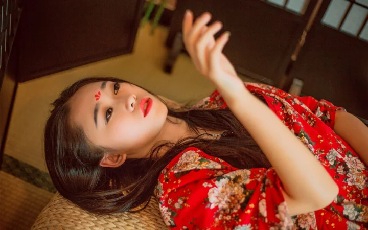 一名成年女性身穿红色花卉服装，侧躺在室内编织垫上并抬手看向镜头。