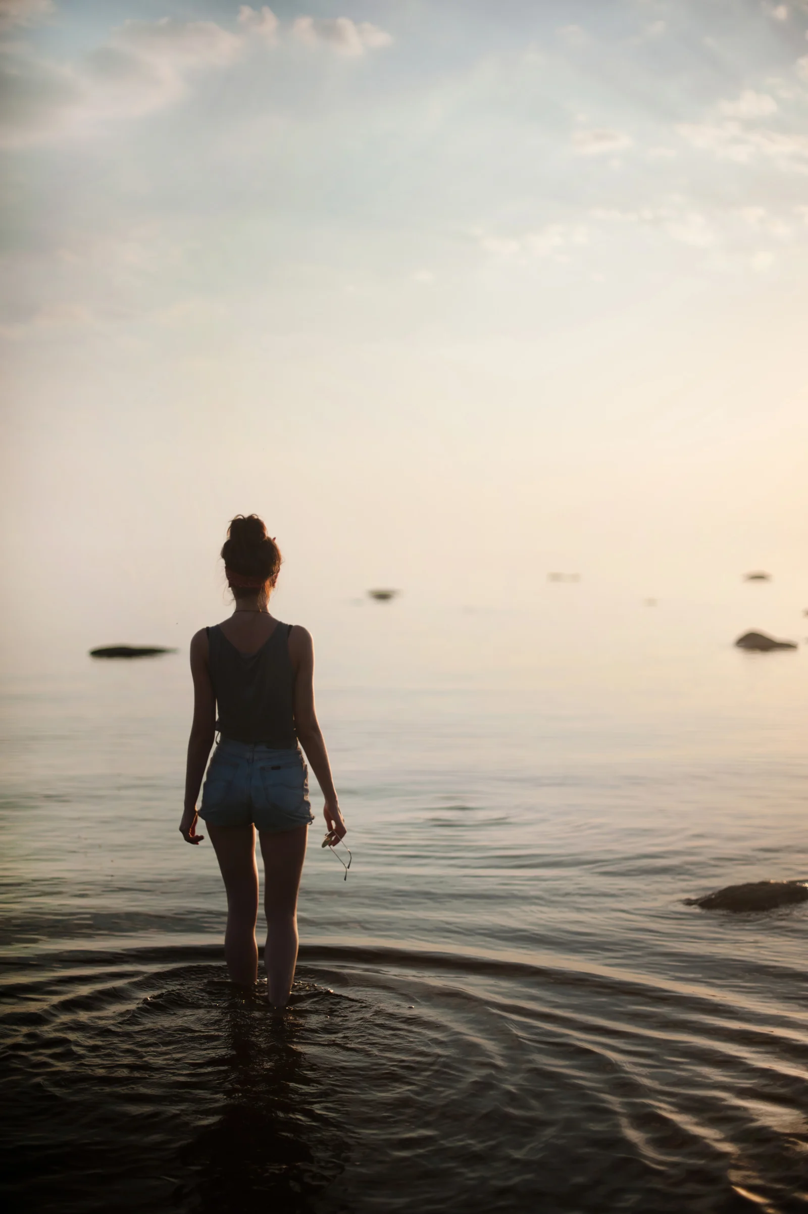 女性が浅瀬に立ち、柔らかな暖色の光に包まれた穏やかな海を見つめている。
