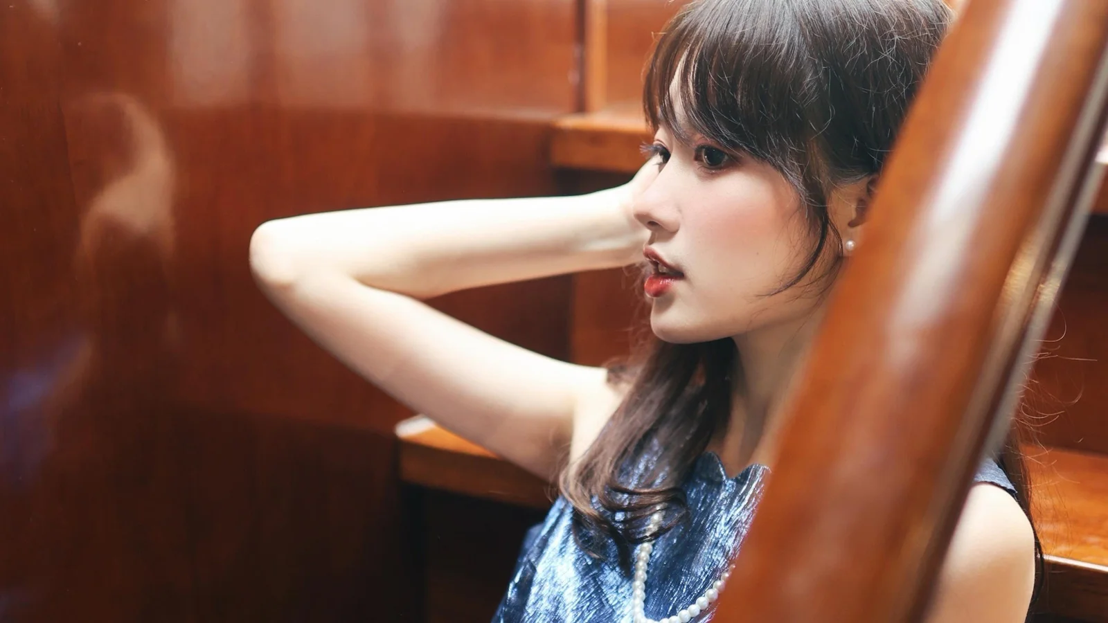 一名成年女性侧身面对镜头，身穿蓝色无袖上衣，置身于木质室内环境中。