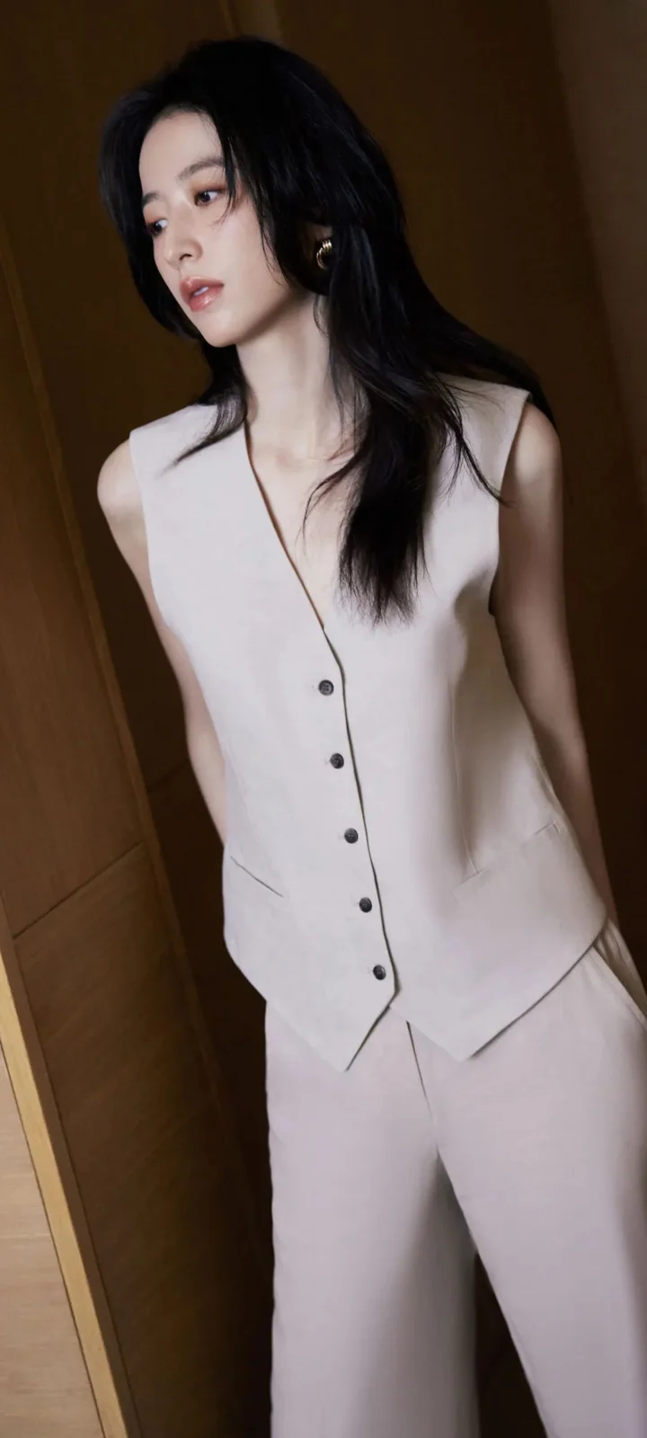 一位成年女性身穿浅色无袖套装站在木质墙面旁。