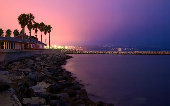 暮色中的海岸边分布着棕榈树、礁石、建筑和远处的灯光，平静海面映出紫粉色天空。