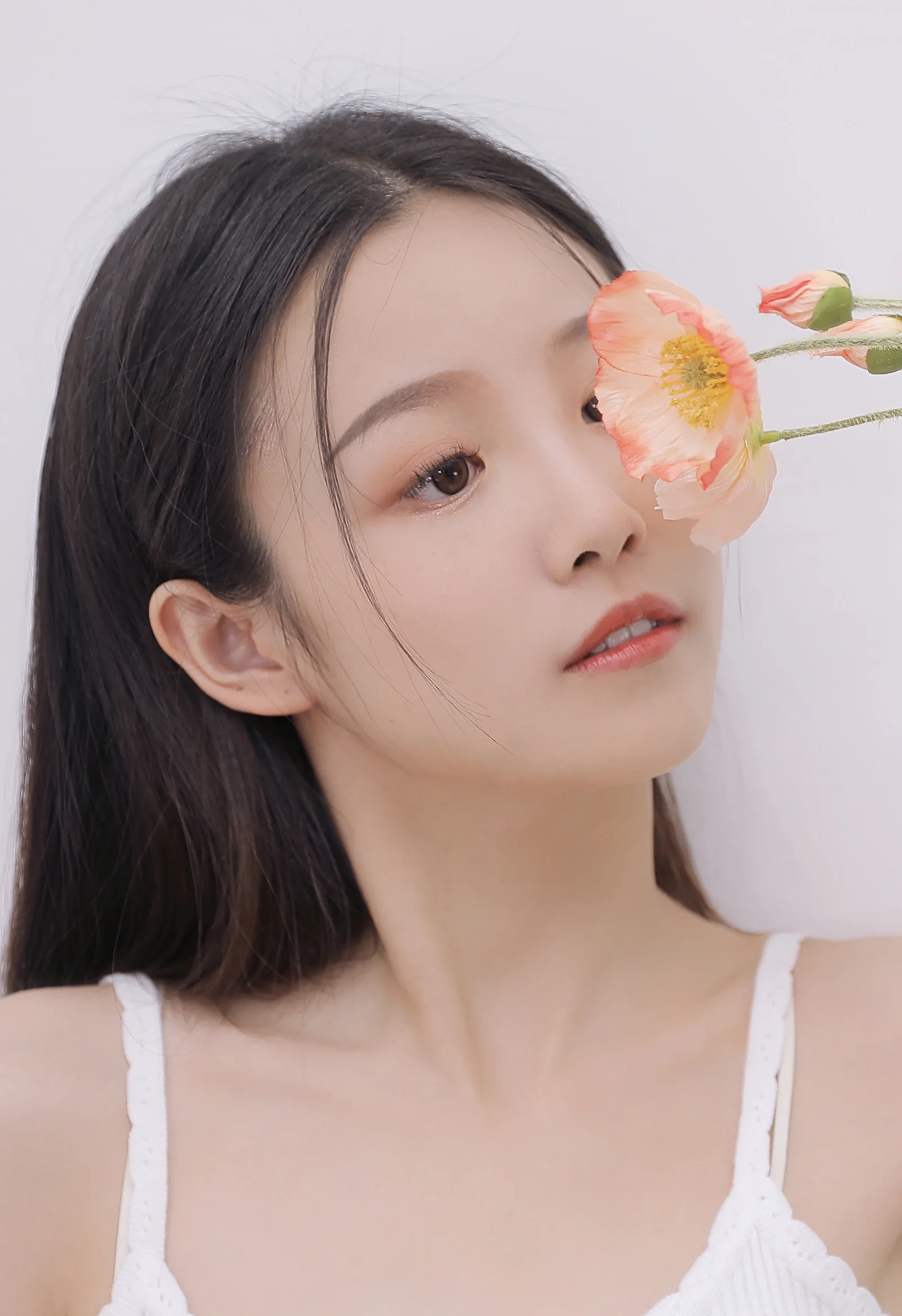 黒髪の女性が、明るくシンプルな背景の中でオレンジピンクの花に顔を寄せています。