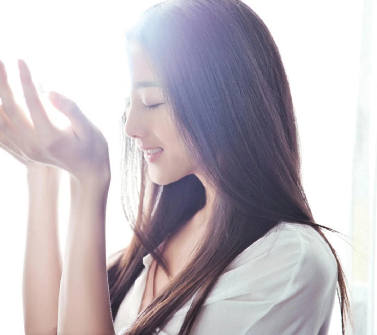 긴 머리의 여성이 밝은 창가에서 눈을 감고 미소 짓고 있다.