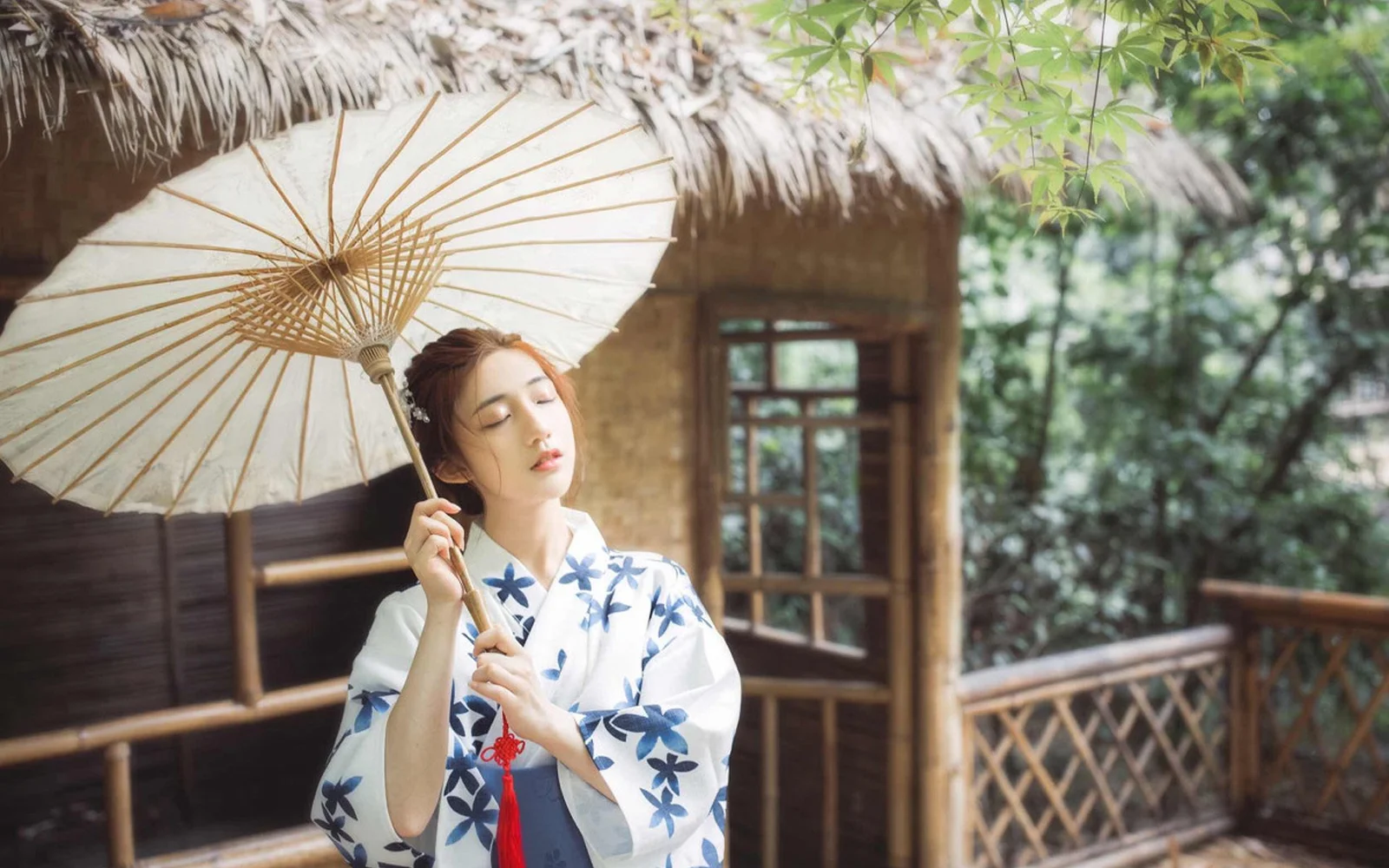 흰 종이 우산을 든 여성이 파란색과 흰색 무늬의 유카타를 입고 목조 건물 앞에 서 있습니다.
