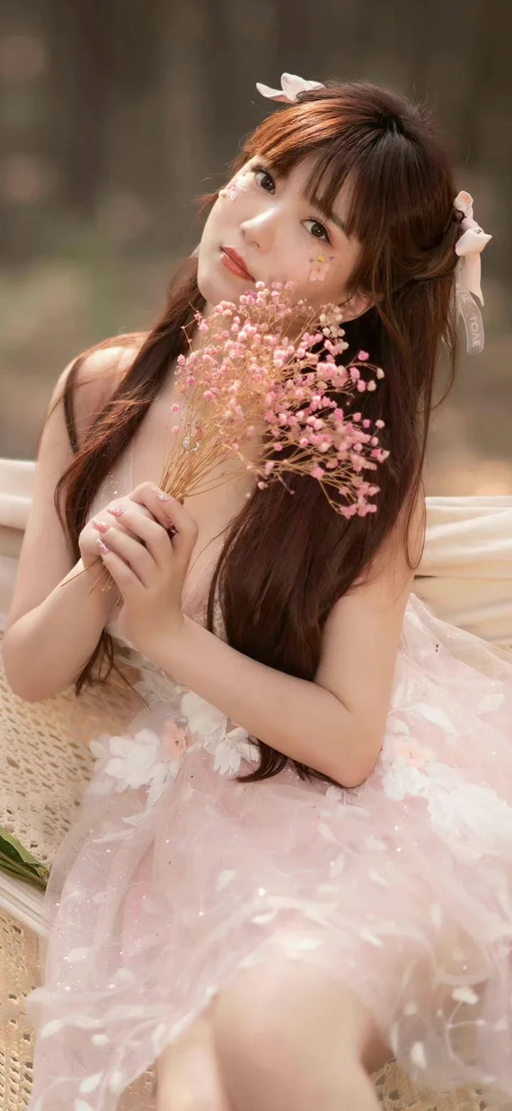 一名棕发女性身穿粉色裙装，手持粉色花束坐在柔和背景前。
