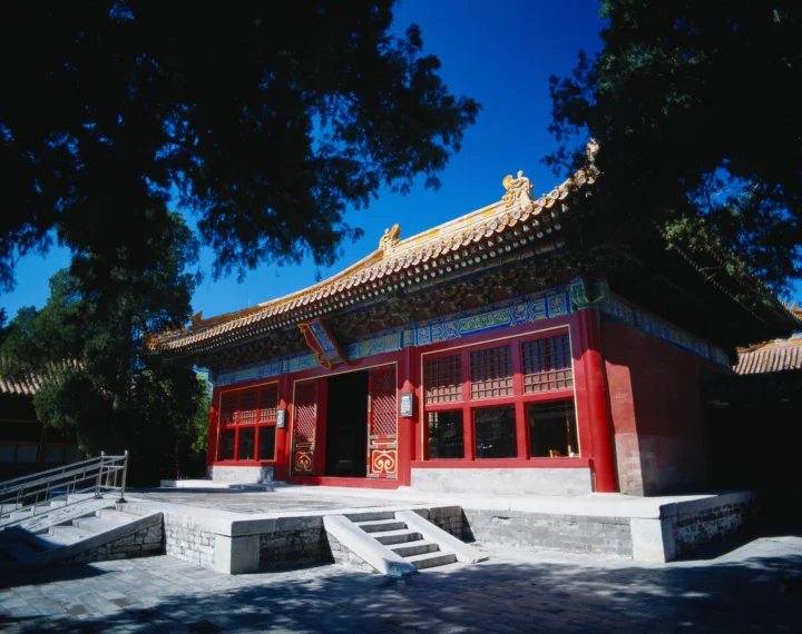 一座红墙彩绘檐饰的中国传统建筑坐落在树木环绕的石阶庭院中。