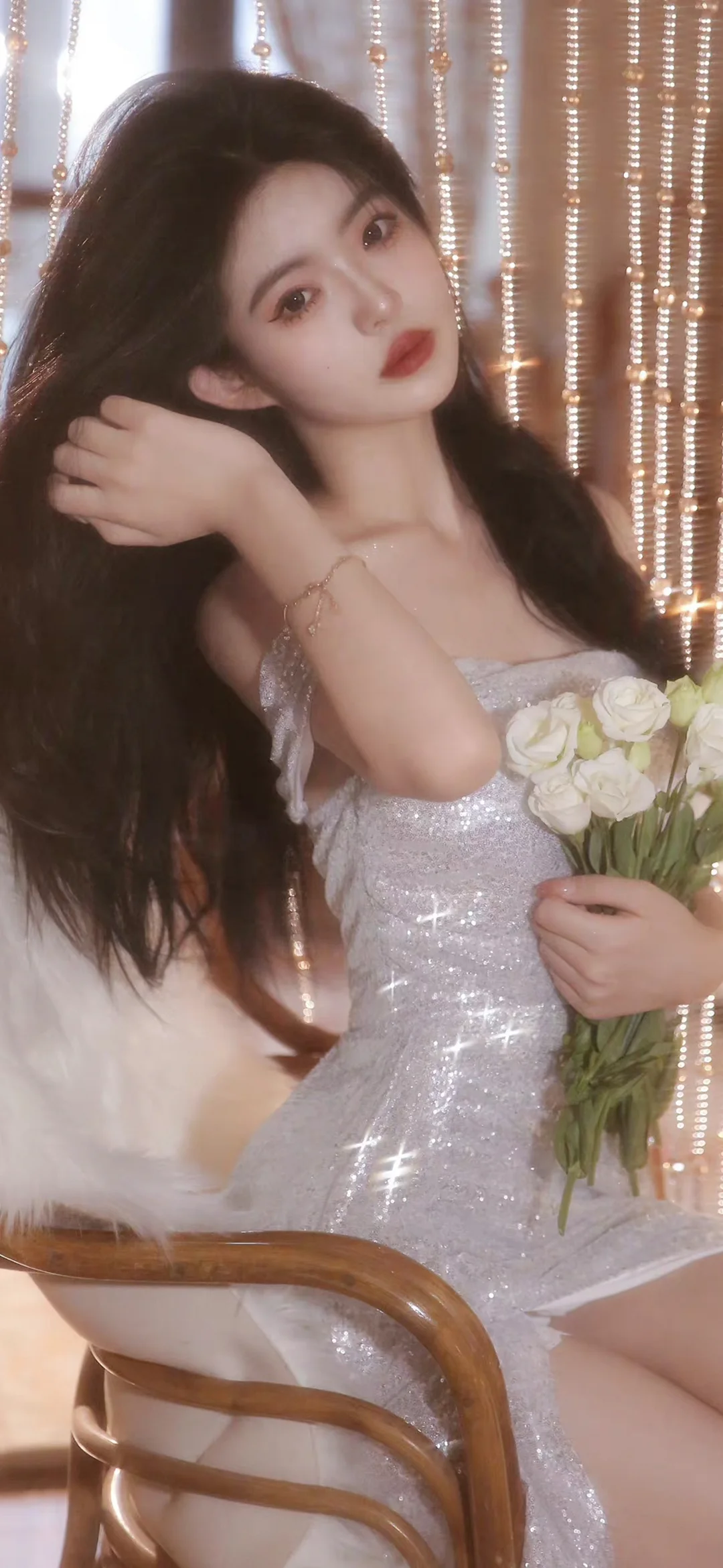 一位长发女性穿着闪亮的银色礼服，手持一束白色花朵坐在室内椅子上。