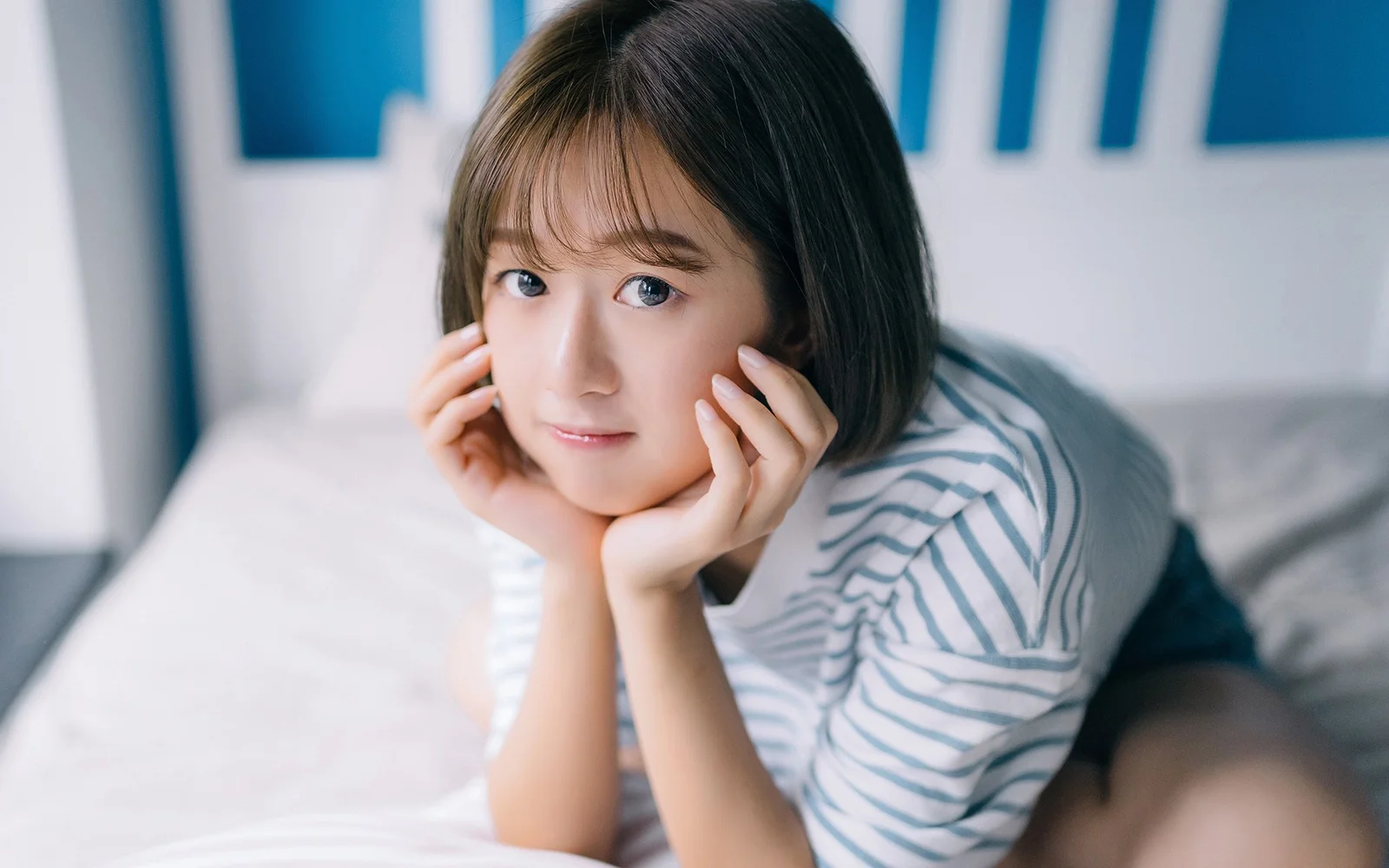一名短发女子穿着条纹上衣趴在床上，用双手托着脸颊看向镜头。