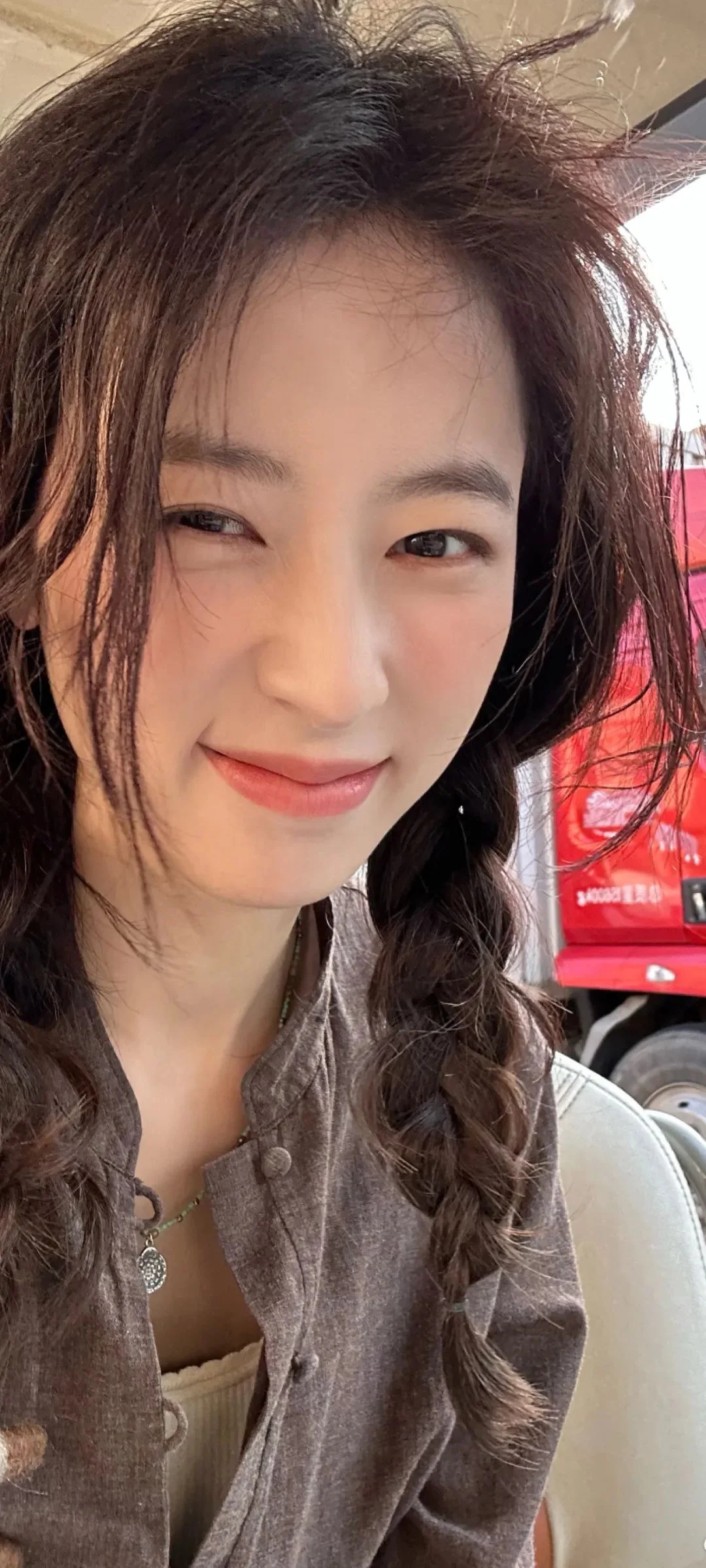 땋은 머리의 여성이 카메라를 향해 미소 짓고 있으며 배경에는 빨간 차량이 보입니다.