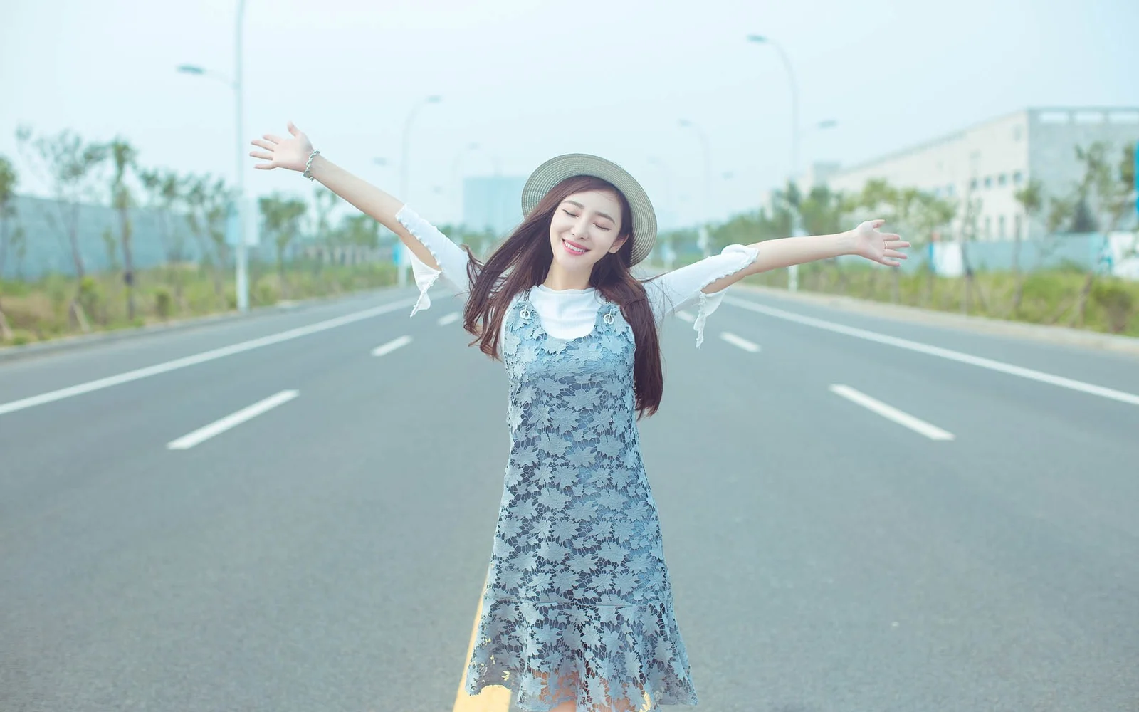 一名戴帽子的年轻女子站在空旷公路中央，张开双臂面向镜头微笑。