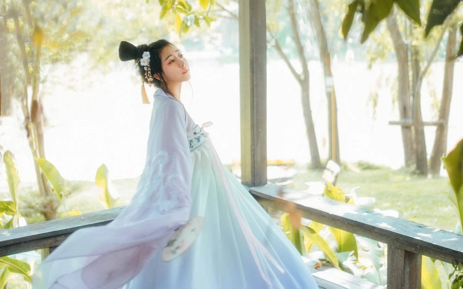 一名身穿浅色传统服饰的女子站在阳光明亮的木质廊台旁，周围可见树木与绿地。