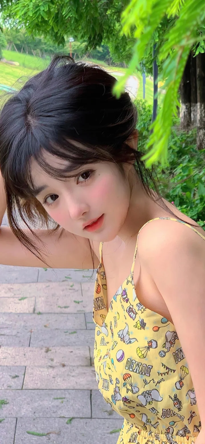 一名黑发女子穿着黄色图案连衣裙，在绿叶环绕的户外步道旁看向镜头。