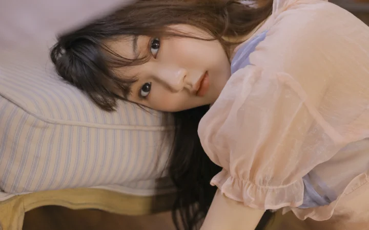 一名长发女性侧躺在床上，面向镜头，穿着浅色泡泡袖上衣。