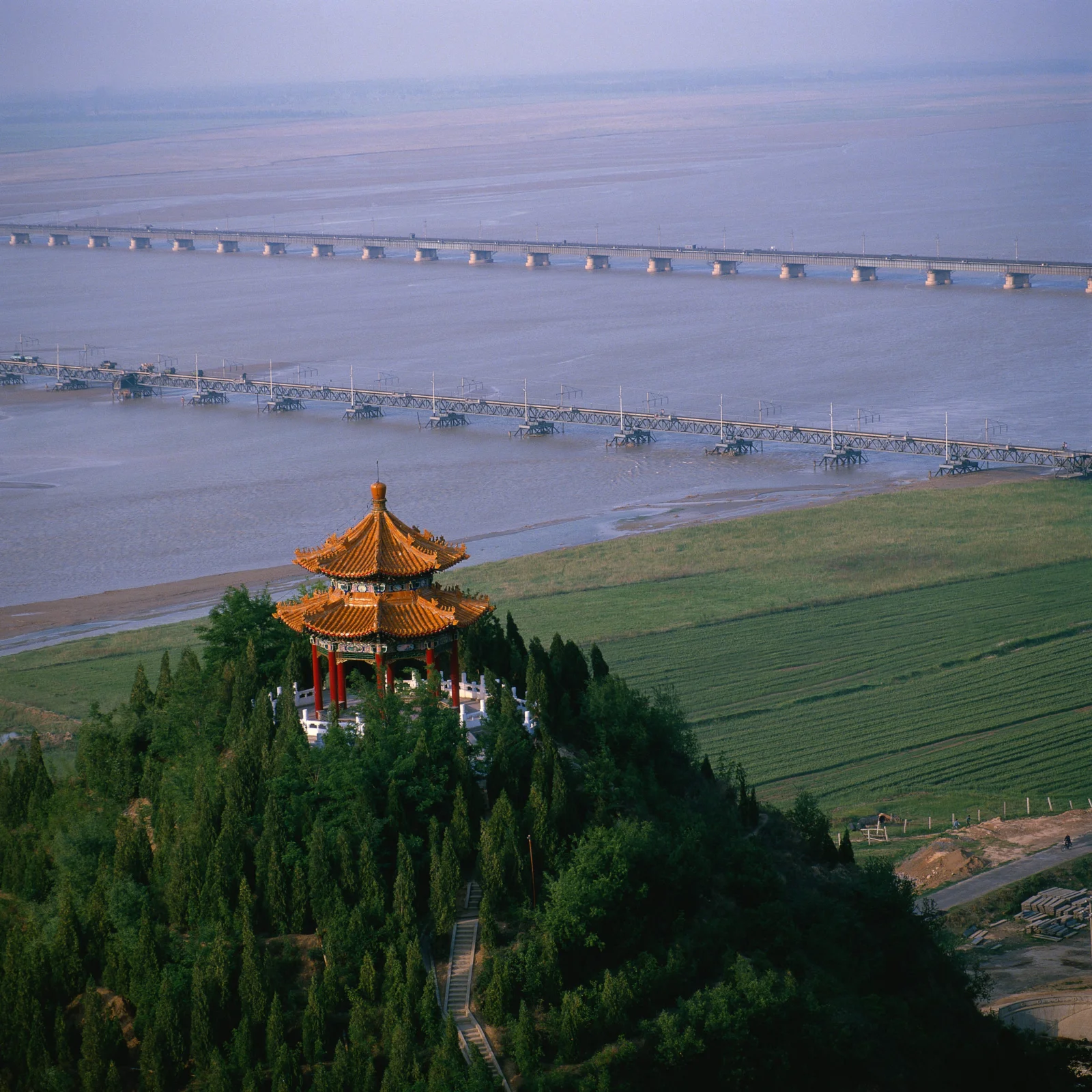 一座传统亭阁坐落在绿树间，远处是宽阔河流、长桥和整齐的农田。